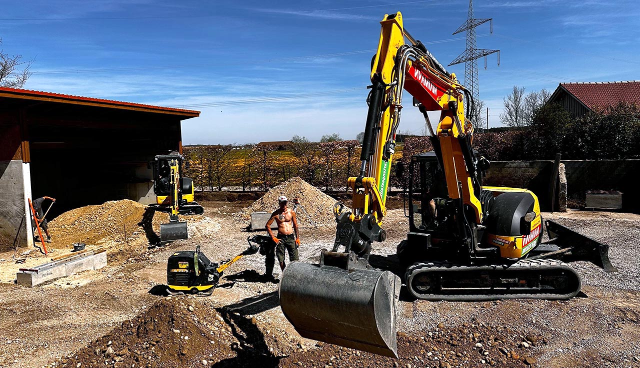 Eine Baustelle von Maimann Gartengestaltung im Landschaftsbau mit zwei gelben Baggern, einer mit angehobener Schaufel. Ein hemdsärmeliger Arbeiter mit Helm steht in der Nähe eines Schotterhaufens. Im Hintergrund sind Gebäude, Bäume und eine Stromleitung unter blauem Himmel zu sehen.