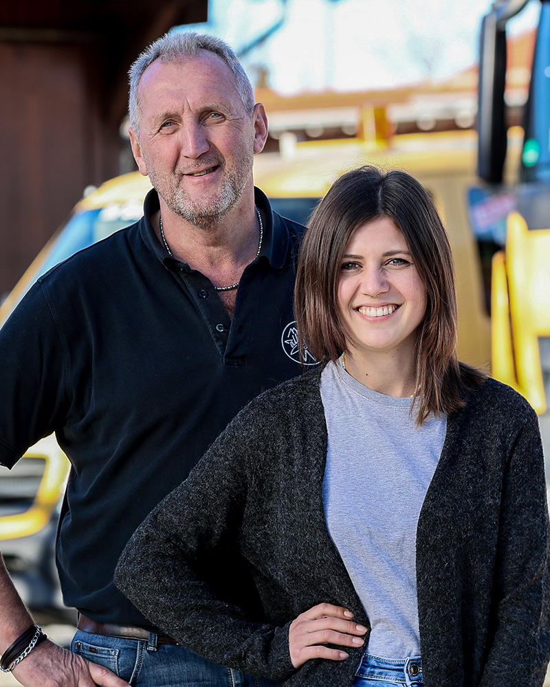 Ein Mann und eine Frau stehen im Freien nebeneinander und lächeln. Der Mann trägt ein schwarzes Poloshirt, die Frau ein graues T-Shirt mit einer dunklen Strickjacke. Im Hintergrund ist ein gelbes Fahrzeug von Maimann Gartengestaltung zu sehen.