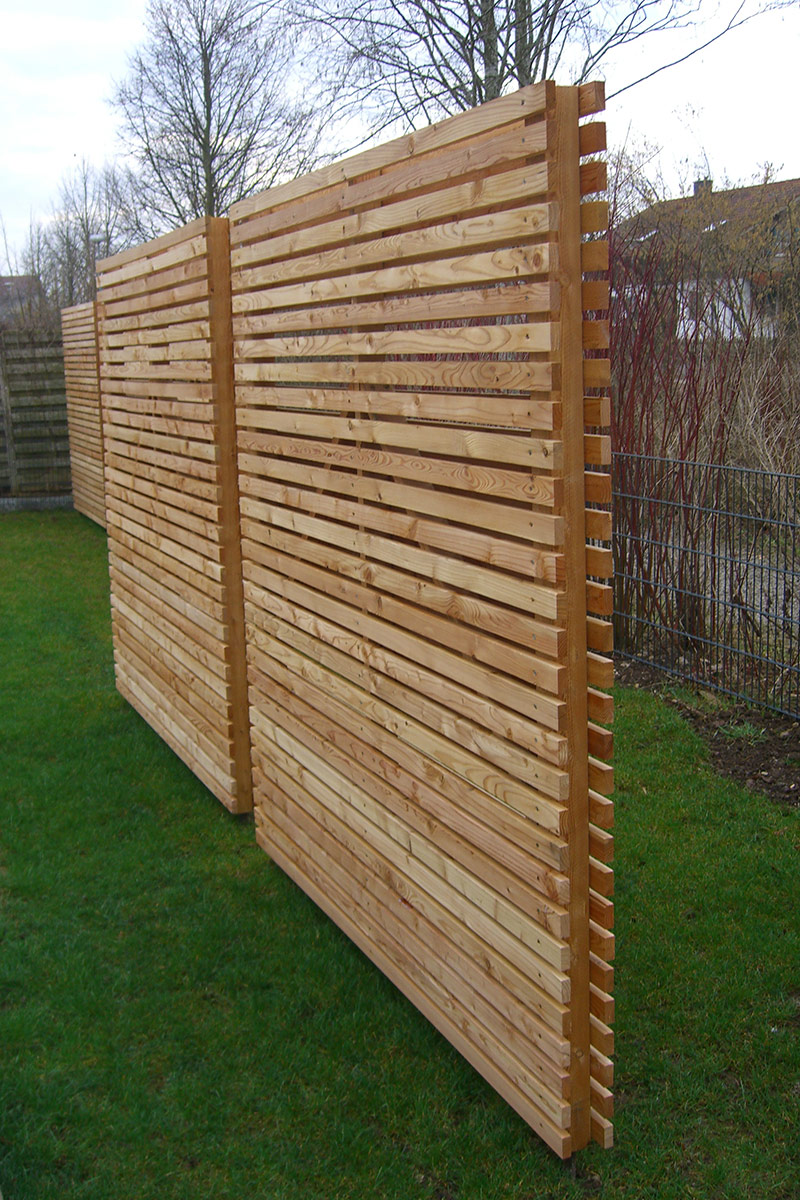 Zwei hohe, moderne Holzzäune mit horizontalen Latten von Maimann Gartengestaltung stehen auf einer grünen Wiese, mit Bäumen und Sträuchern im Hintergrund. Die hellbraunen Zäune scheinen neu errichtet worden zu sein.