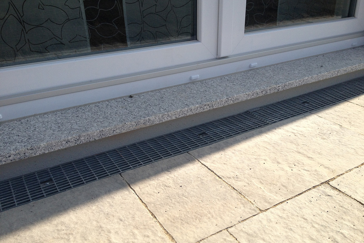 Eine Nahaufnahme der Außenansicht eines Gebäudes von Maimann Gartengestaltung zeigt eine weiß gerahmte Glastür, eine steinerne Fensterbank, ein metallenes Abflussgitter und helle Steinplatten.