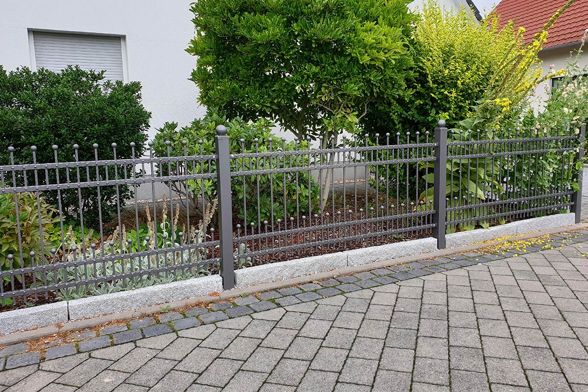 Ein Metallzaun mit vertikalen Stäben steht vor einem von Maimann Gartengestaltung gestalteten Garten mit grünen Sträuchern und einem Baum, der von einem gepflasterten Gehweg begrenzt wird. Im Hintergrund ist ein weißes Gebäude mit einem Fenster zu sehen.