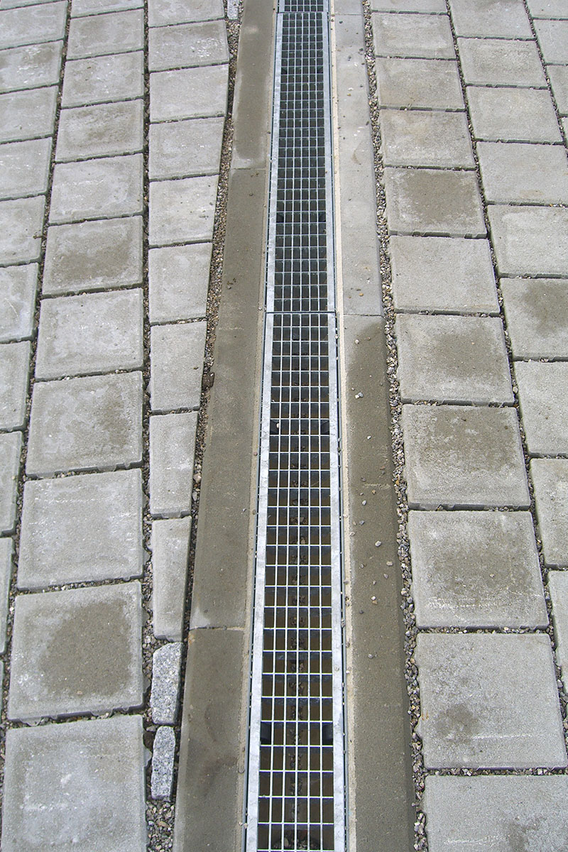 Ein Metallgitter bedeckt eine lange, schmale Entwässerungsrinne, die zwischen Reihen von rechteckigen Betonpflastersteinen auf einer gepflasterten Fläche verläuft und die für die Maimann Gartengestaltung typische präzise Gestaltung zeigt.