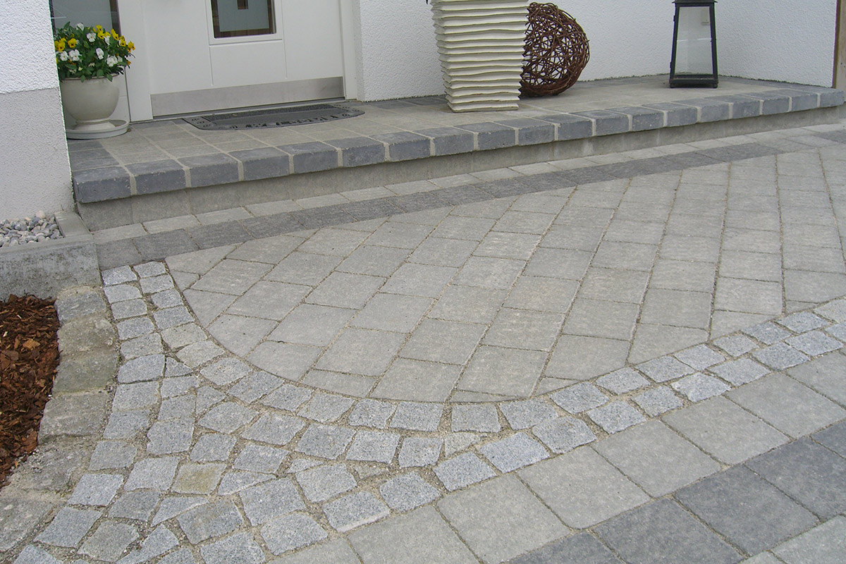 Ein moderner Hauseingang von Maimann Gartengestaltung, mit einem gemusterten Gehweg aus grauen Pflastersteinen in geschwungenen und geraden Linien, einer Willkommensmatte, Topfblumen, einem großen Pflanzgefäß und einer dekorativen Laterne.