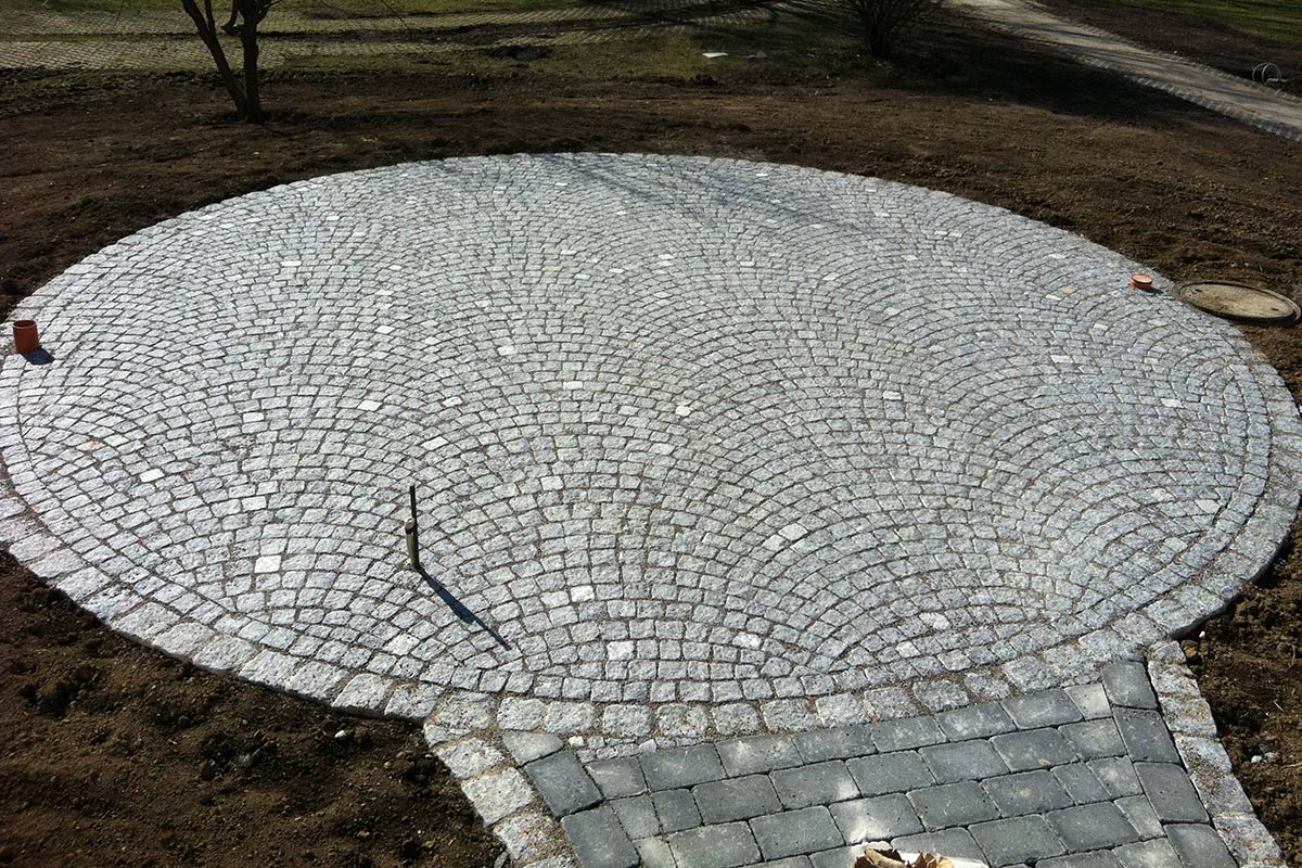 Eine kreisförmige Steinterrasse von Maimann Gartengestaltung aus kleinen, hellgrauen Pflastersteinen in Fächermuster mit einem kurzen, rechteckigen Weg, der vom unteren Rand aus zu ihr führt, ist in nackte Erde eingelassen.