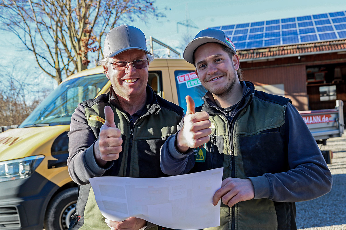 Zwei Männer mit Mützen und Arbeitsjacken stehen im Freien, lächeln und zeigen die Daumen nach oben, während sie einen Satz Pläne halten. Hinter ihnen steht ein gelber Lieferwagen mit dem Logo von Maimann Gartengestaltung und ein Gebäude mit Solarzellen auf dem Dach.