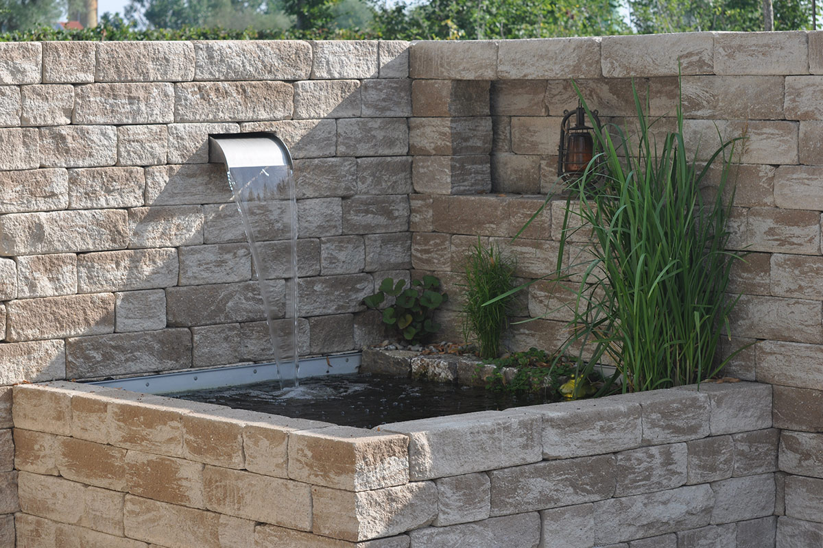 Ein von Maimann Gartengestaltung angelegter Teich mit einem steinernen Wasserfall, Wasserfontänen, üppigem Gras, blühenden Sträuchern und einer gemauerten Sichtschutzmauer lädt zum Verweilen im Freien ein.