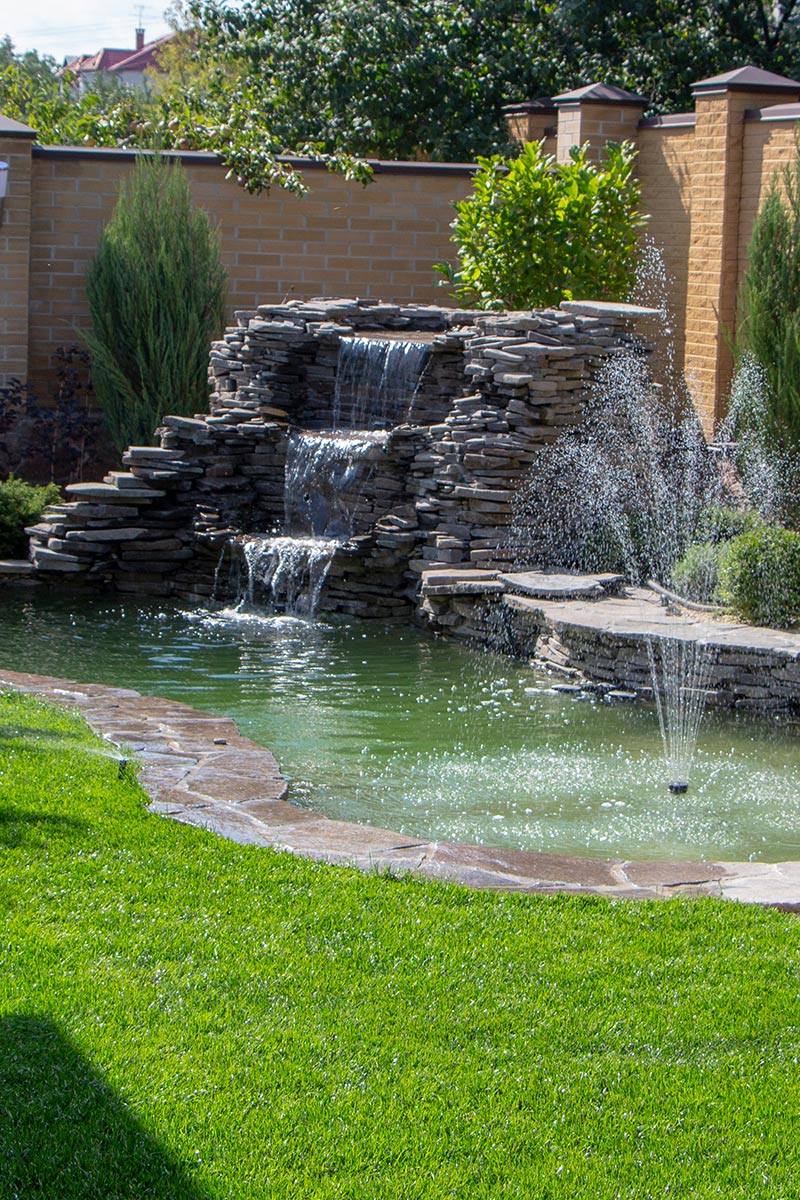 Ein Hinterhof mit einem kleinen steinernen Wasserfall, der in einen Teich fließt, entworfen von Maimann Gartengestaltung; ein Springbrunnen spritzt Wasser in den Teich, umgeben von grünem Gras und ordentlich gestutzten Büschen, mit einer Ziegelmauer im Hintergrund.