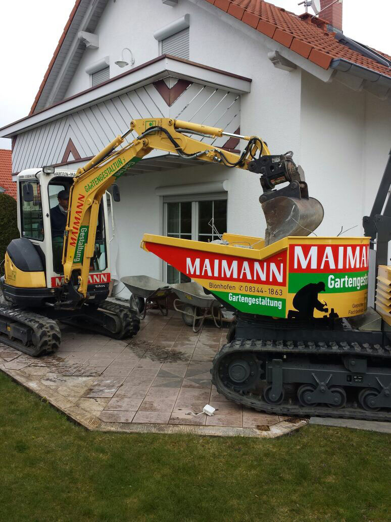 Ein gelber Bagger von Maimann Gartengestaltung wirft Schutt in eine rot-gelbe Mulde auf einer gefliesten Terrasse vor einem weißen Haus ab. Eine andere Maschine hält die Mulde, während beide in der Nähe des Hauses geparkt sind.
