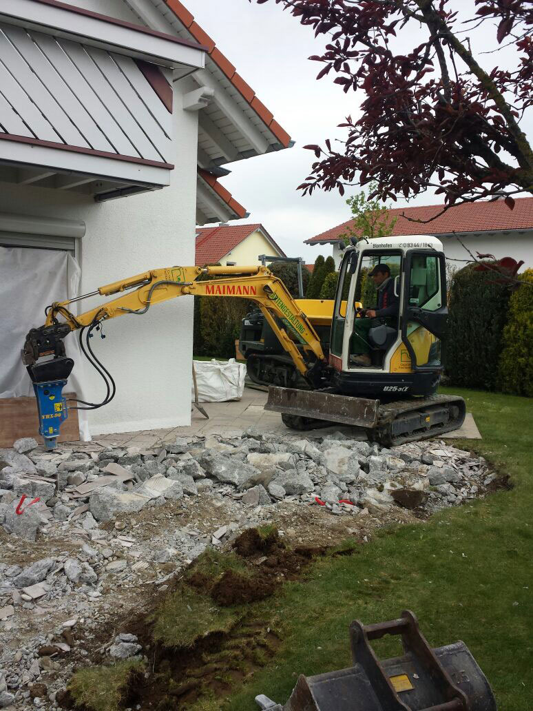 Ein gelber Minibagger von Maimann Gartengestaltung bricht in der Nähe eines weißen Hauses Beton ab, wobei Schutt auf dem Boden verstreut ist und rechts ein Baum teilweise sichtbar ist.