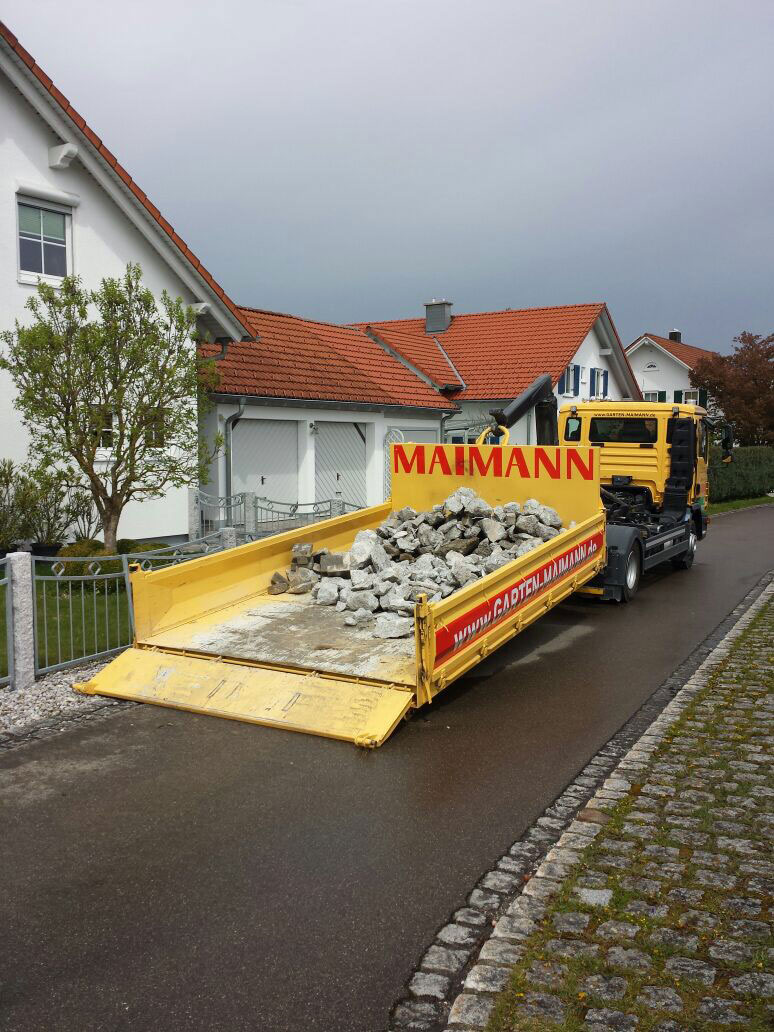 Ein gelber Lkw mit kippbarer Ladefläche von Maimann Gartengestaltung ist in einer Wohnstraße geparkt und trägt eine Ladung zerbrochenen Betonschutts. Der Lkw steht vor weißen Häusern mit roten Dächern und einem gepflegten Bürgersteig.