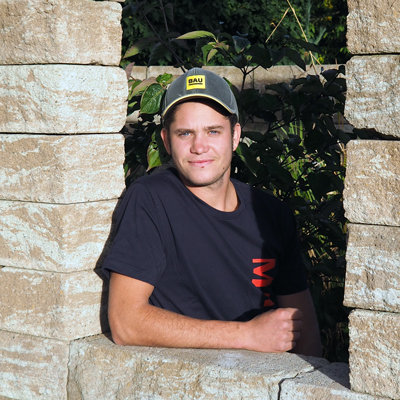 Christoph Berg mit schwarzem T-Shirt und dunkler Baseballmütze lehnt an einer Steinmauer im Freien. Im Hintergrund ist üppiges Grün zu sehen, das möglicherweise von Maimann Gartengestaltung entworfen wurde, und das Sonnenlicht beleuchtet sein Gesicht.