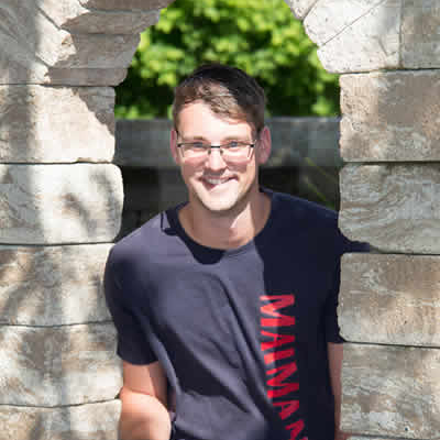 Domenik Prestele mit Brille und marineblauem T-Shirt steht lächelnd in einem steinernen Torbogen, das Sonnenlicht beleuchtet sein Gesicht und im Hintergrund ist das Grün der Maimann Gartengestaltung zu sehen.