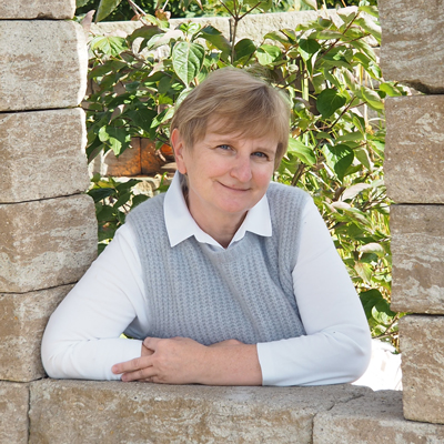 Karin Walcz mit kurzen blonden Haaren, die ein weißes Hemd und eine graue Weste trägt, lehnt an einer Steinmauer mit üppigem Grün im Hintergrund und lächelt in die Kamera - eine Anspielung auf den Stil von Maimann Gartengestaltung.