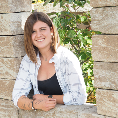 Lena Maimann mit schulterlangen braunen Haaren, die ein schwarzes Top und ein weißes kariertes Hemd trägt, lehnt lächelnd an einer von Maimann Gartengestaltung entworfenen Steinmauer mit grünen Blättern im Hintergrund.