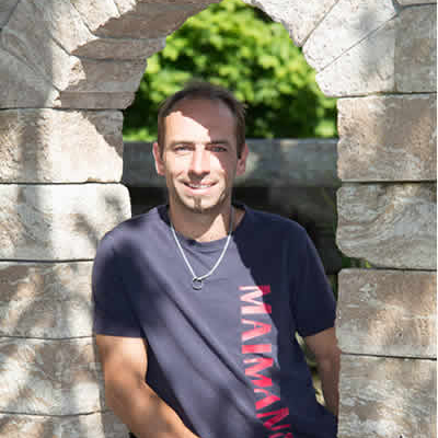 Michael Zaumseil mit kurzen Haaren und Bart, der ein marineblaues T-Shirt und eine Halskette trägt, sitzt in einem steinernen Torbogen, der von Maimann Gartengestaltung gefertigt wurde, mit Sonnenlicht und Grün im Hintergrund.