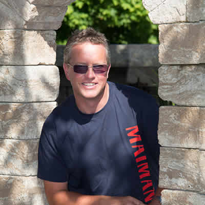 Rudolf Glöckler, der eine Sonnenbrille und ein marineblaues T-Shirt mit roter Aufschrift trägt, steht lächelnd zwischen zwei Steinsäulen, im Hintergrund eine üppige Grünanlage von Maimann Gartengestaltung.